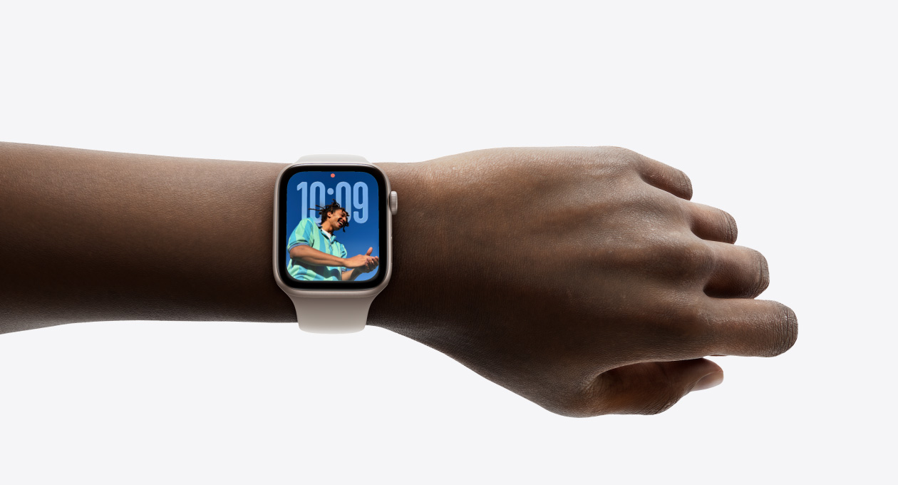 Primer plano de un Apple Watch en oro rosa con correa de goma en la muñeca de una persona que muestra la hora con el nuevo diseño en Liquid Glass
