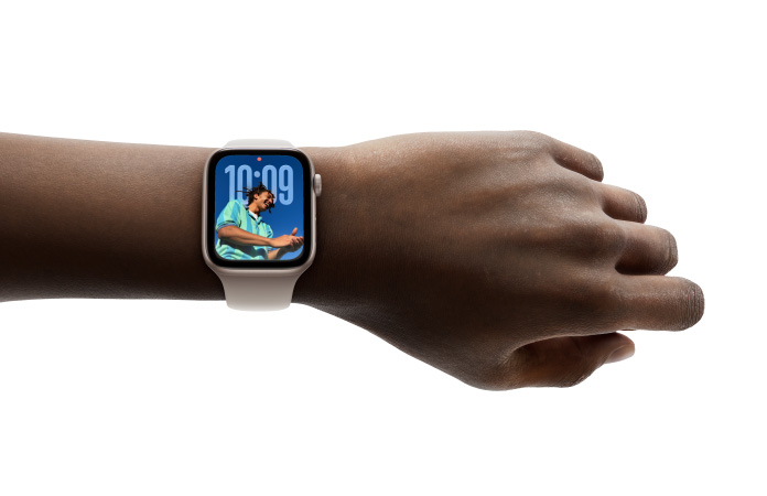 Grande plano do Apple Watch em rosa‑dourado com bracelete de borracha no pulso, a mostrar a app Relógio no novo design com Liquid Glass