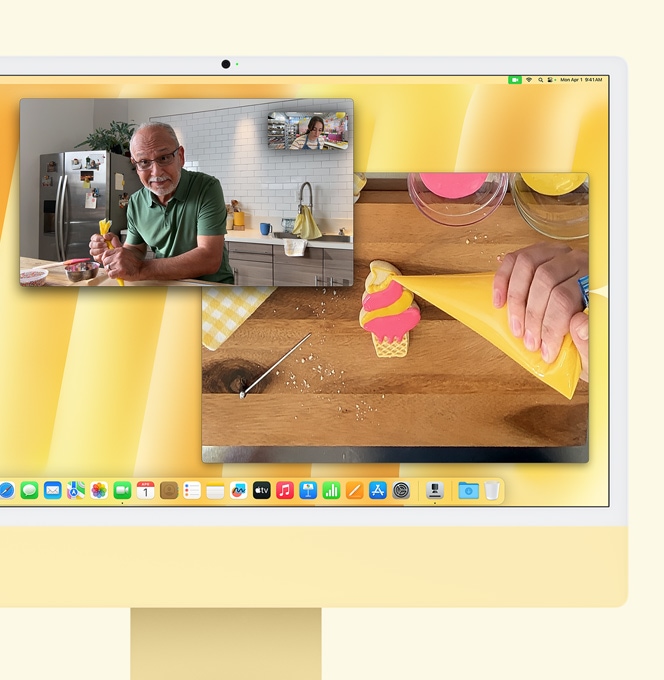 Egy sárga iMac az Íróasztal nézet működését szemlélteti. A kijelzőn egy videohívás látható, amelyben egy cukrász egy másik emberrel beszélget. Az egyik videóablak a beszélgetőtársat és a cukrászt mutatja szemből, egy másikban pedig a cukrász keze látszik felülnézetből, ahogy cukormázzal díszít egy kekszet.