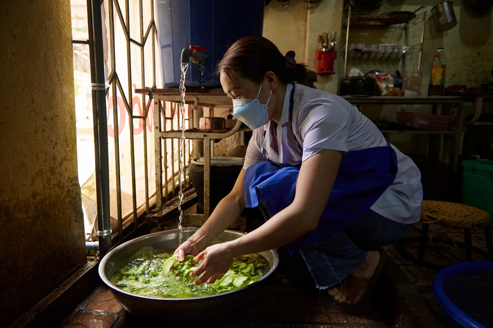 Una persona con mascarilla y delantal lava verduras en un gran recipiente con agua que sale de una llave.