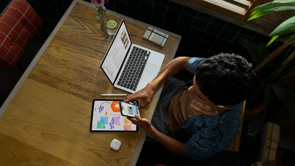 An overhead view shows a person holding iPhone and working at a table with MacBook Pro, which features coding on the screen, alongside iPad, Apple Pencil, and AirPods.