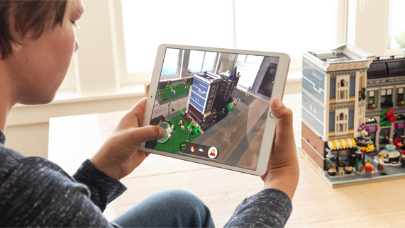Un garçon assis devant une construction en LEGO dans une pièce, tenant un iPad sur l’écran duquel s’affiche LEGO AR City. 