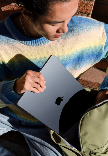 一名學生坐在户外。他從背囊中拿出一台合上的 MacBook。