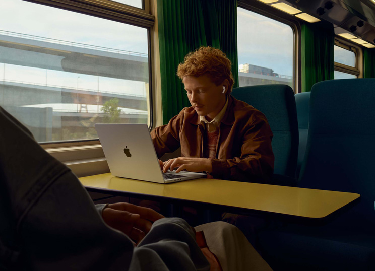 Une personne dans un train, assise près d’une fenêtre, utilise des AirPods et travaille sur un MacBook.