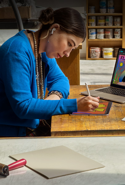 Two students view content on a silver MacBook in their dorm room, with one sitting on the bed as the other sits on the floor holding the device