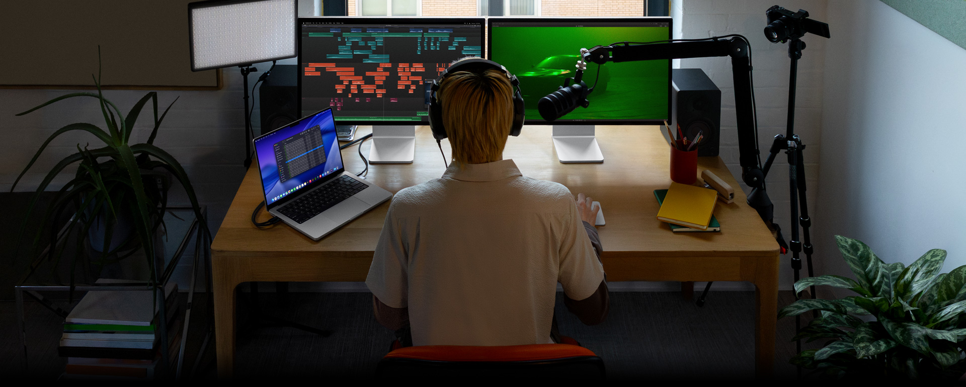 Eine Person sitzt an einem Schreibtisch mit Mikrofon und Kopfhörern vor drei Geräten, ein Mac Studio Bildschirm zeigt detaillierte Tools für die Audiobearbeitung mit Soundclips in verschiedenen Farbblöcken, ein zweiter Mac Studio Bildschirm zeigt ein Auto in stimmungsvollem grünen Licht, ein 16 Zoll MacBook Pro Bildschirm zeigt eine lange Liste von Aufnahmen in einem Final Cut Pro Projekt