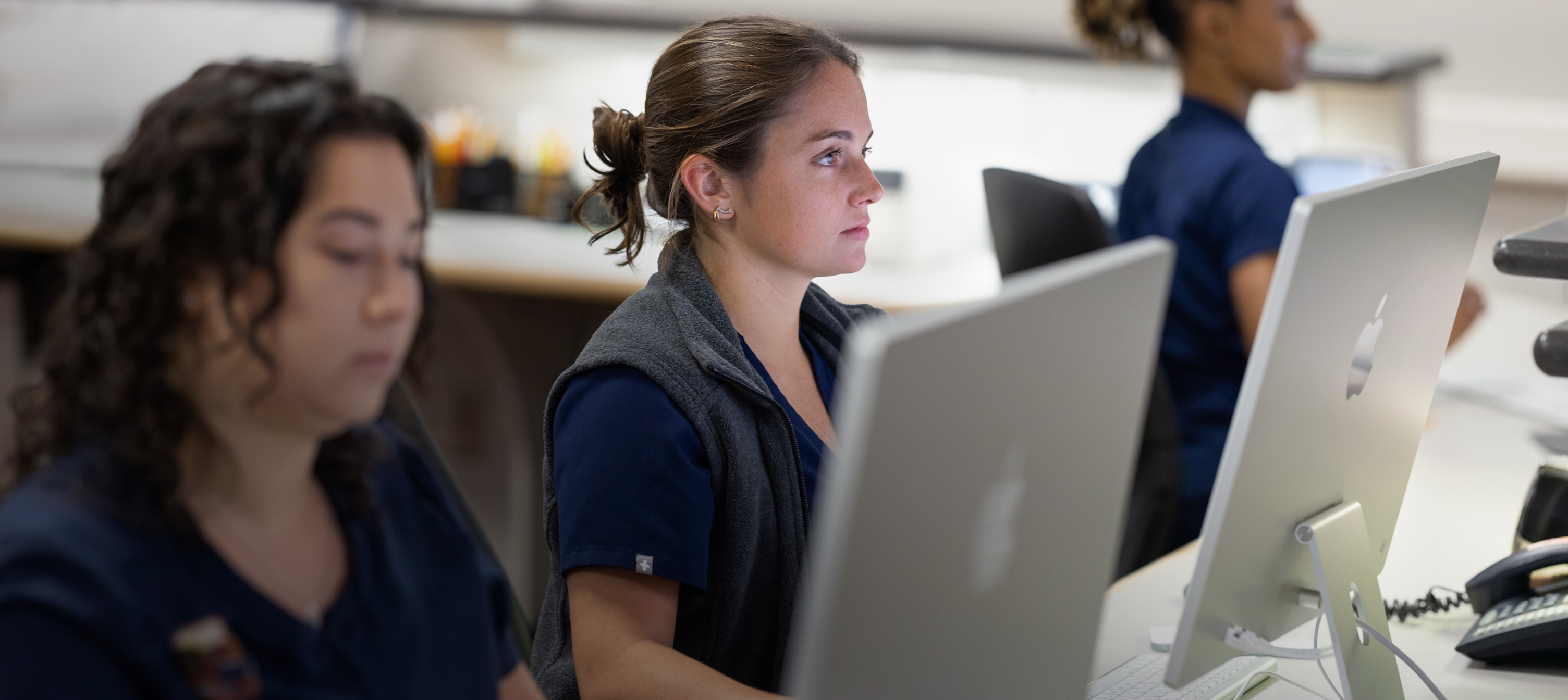 Tre sykepleiere i arbeid på iMac-skjermer.