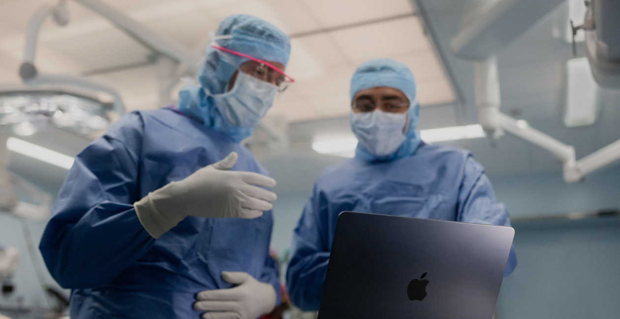 To kirurger diskuterer noe på en MacBook Pro-skjerm inne på operasjonssalen.
