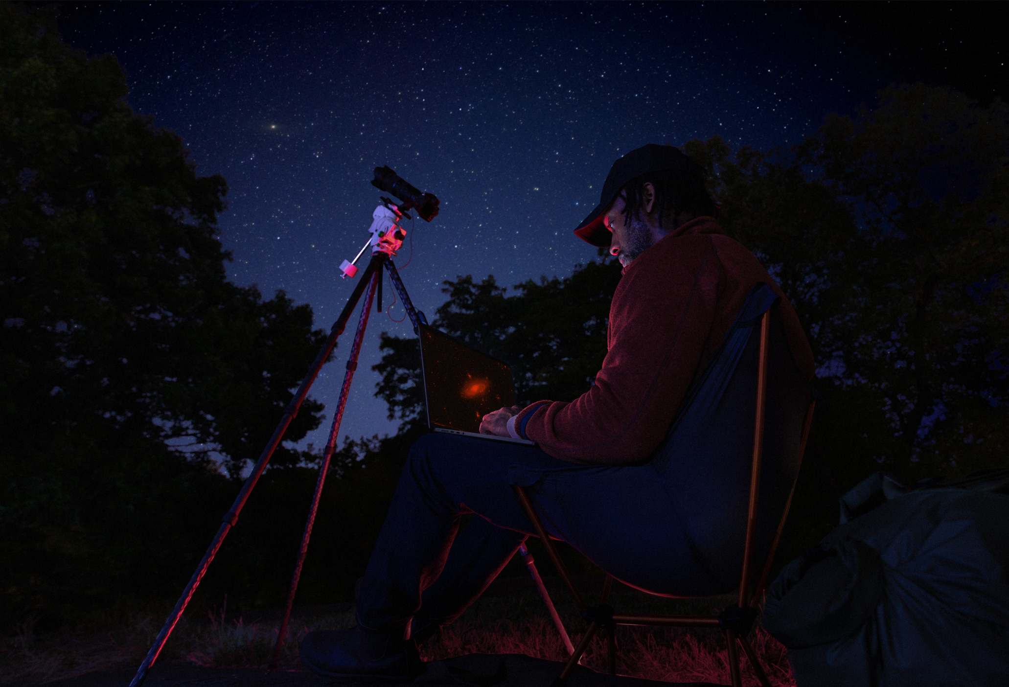 Una persona usa su MacBook Pro sobre el regazo de noche al aire libre, conectado a un dispositivo sobre un trípode que apunta al cielo estrellado